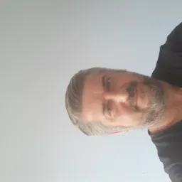 Portret brodatego mężczyzny z siwymi włosami, ubranego w czarną koszulkę, na jasnym tle.