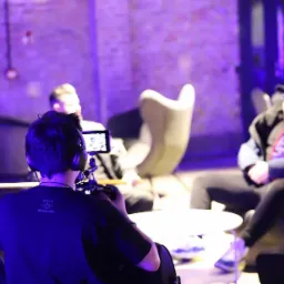 Operator kamery z tyłu, w słuchawkach, nagrywa wywiad z dwoma osobami siedzącymi w designerskich fotelach przy okrągłym stoliku, w pomieszczeniu oświetlonym fioletowym światłem.