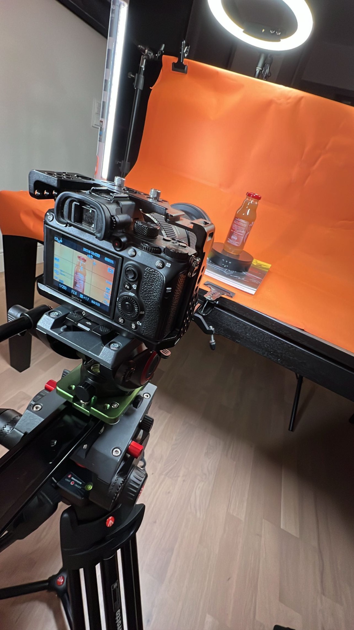 Profesjonalny aparat na statywie nagrywający butelkę napoju na obrotowej platformie na tle pomarańczowego ekranu, oświetlony lampą pierścieniową i listwą LED.