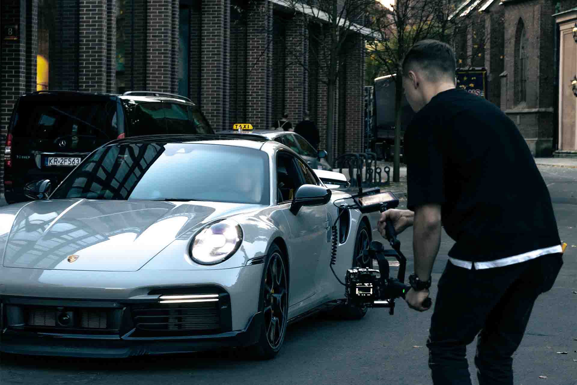 Filmowiec nagrywa ujęcia srebrnego Porsche 911 na tle miejskiej ulicy z budynkami z czerwonej cegły i zaparkowaną taksówką.