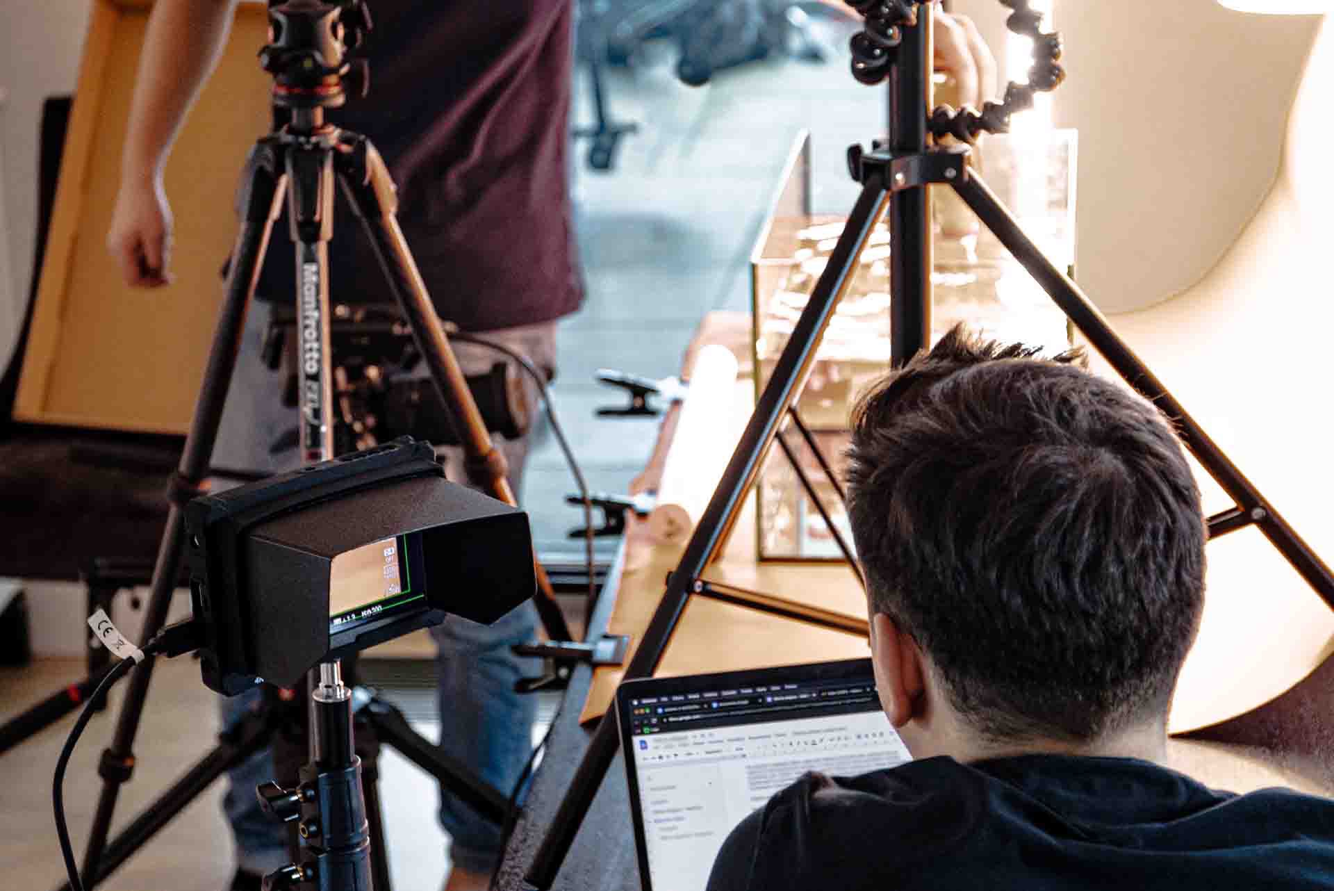 Praca nad montażem filmu: mężczyzna siedzi przed laptopem, obok sprzęt oświetleniowy i kamera na statywie z zamontowanym monitorem podglądowym.