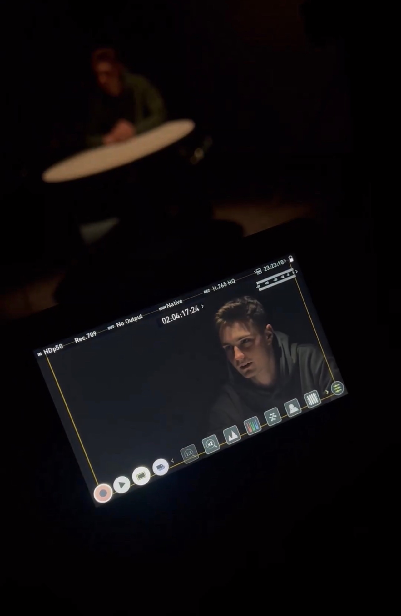 Ekran monitora podglądowego z młodym mężczyzną w trakcie nagrywania filmu, w tle zamazana sylwetka osoby siedzącej przy stole.