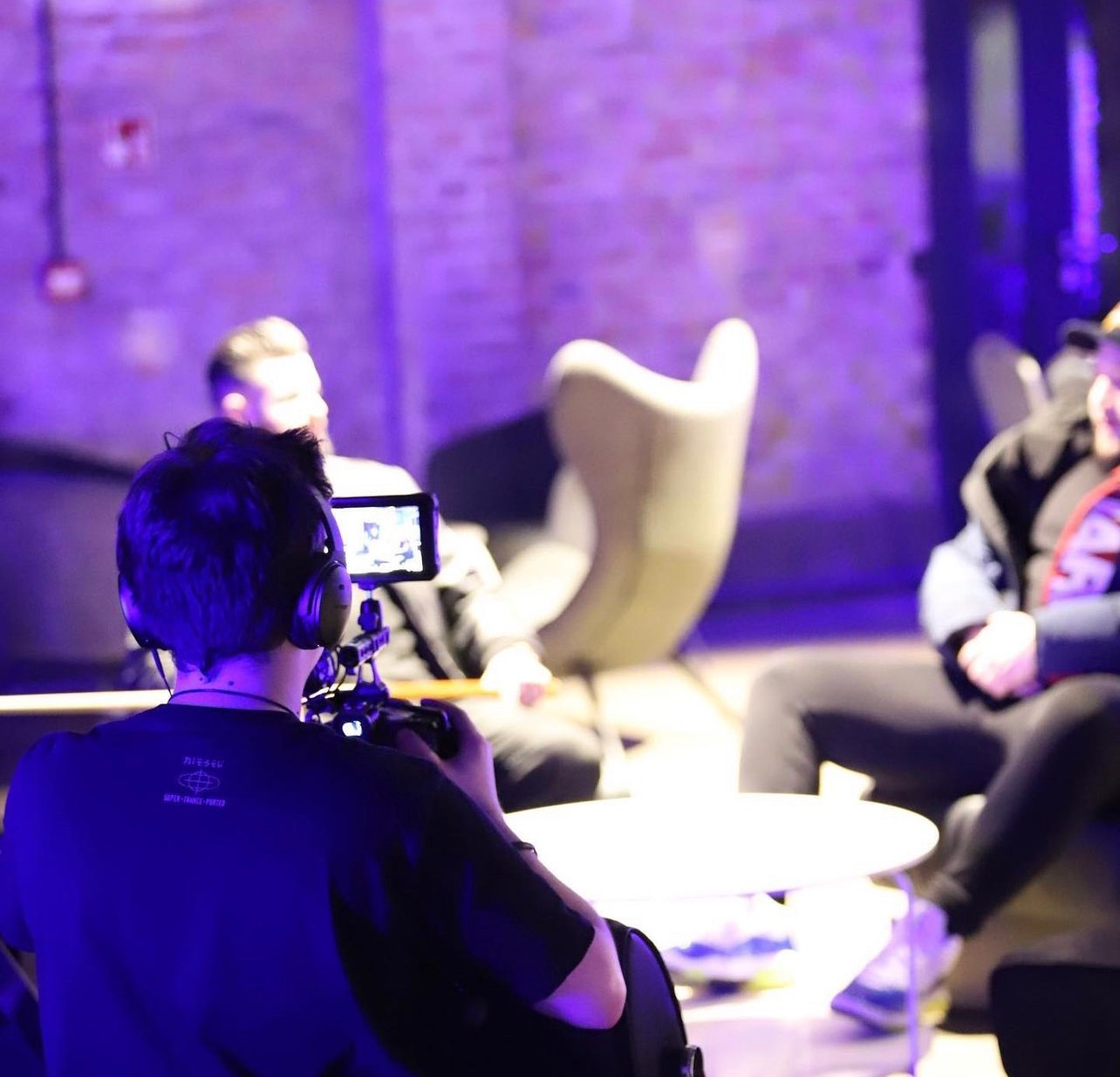 Operator kamery z tyłu, w słuchawkach, nagrywa wywiad z dwoma osobami siedzącymi w designerskich fotelach przy okrągłym stoliku, w pomieszczeniu oświetlonym fioletowym światłem.