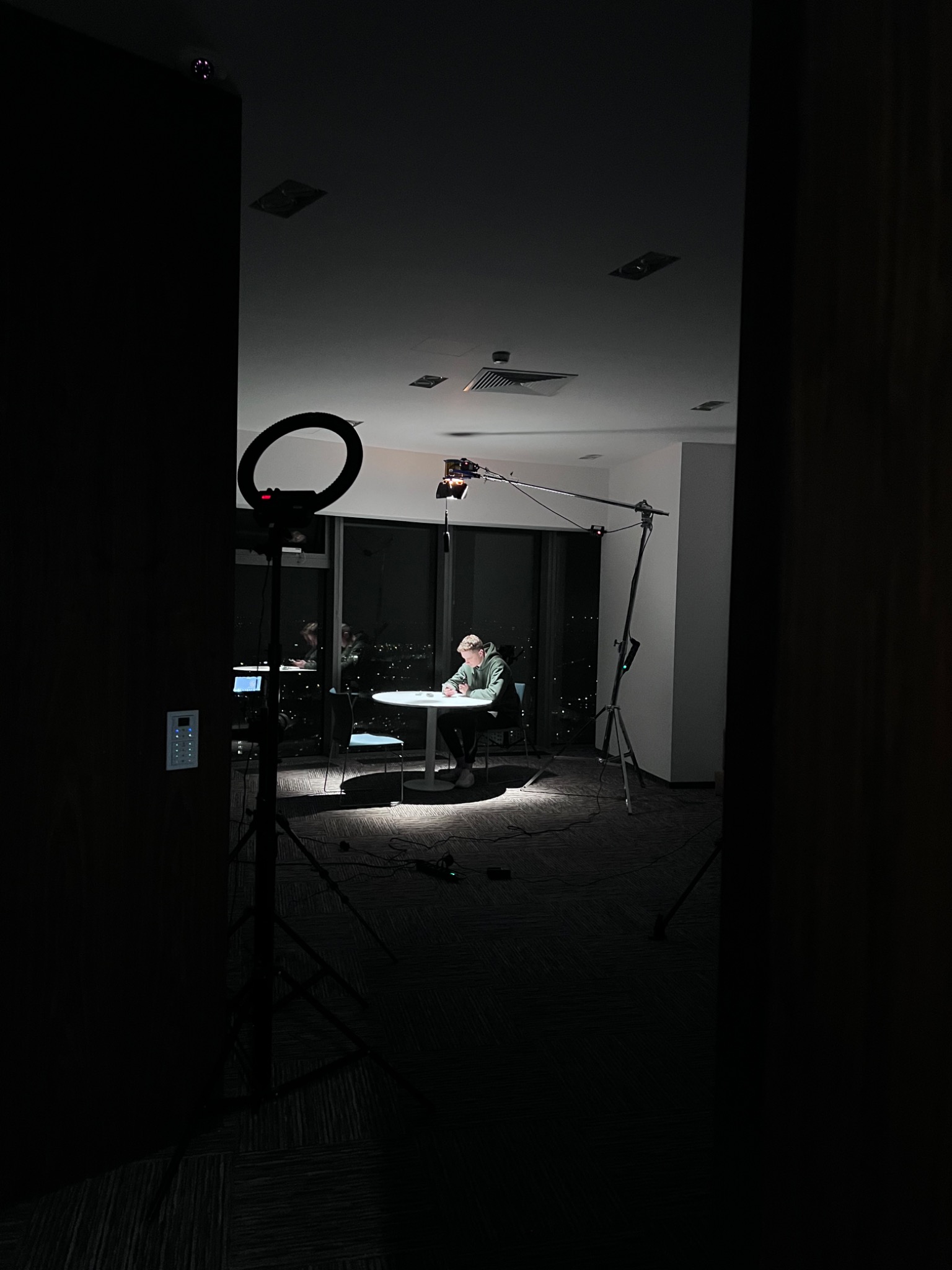 Osoba siedząca przy okrągłym stole w oświetlonym pomieszczeniu, otoczona sprzętem do nagrywania wideo, w tym lampą pierścieniową i mikrofonem na wysięgniku.