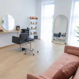 Jasne wnętrze salonu z dwoma fotelami fryzjerskimi, lustrami w złotych ramach, regałem z kosmetykami i różową sofą dla klientów.