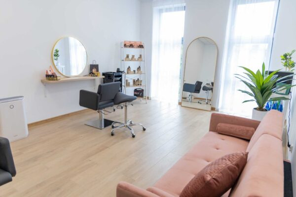 Jasne wnętrze salonu z dwoma fotelami fryzjerskimi, lustrami w złotych ramach, regałem z kosmetykami i różową sofą dla klientów.