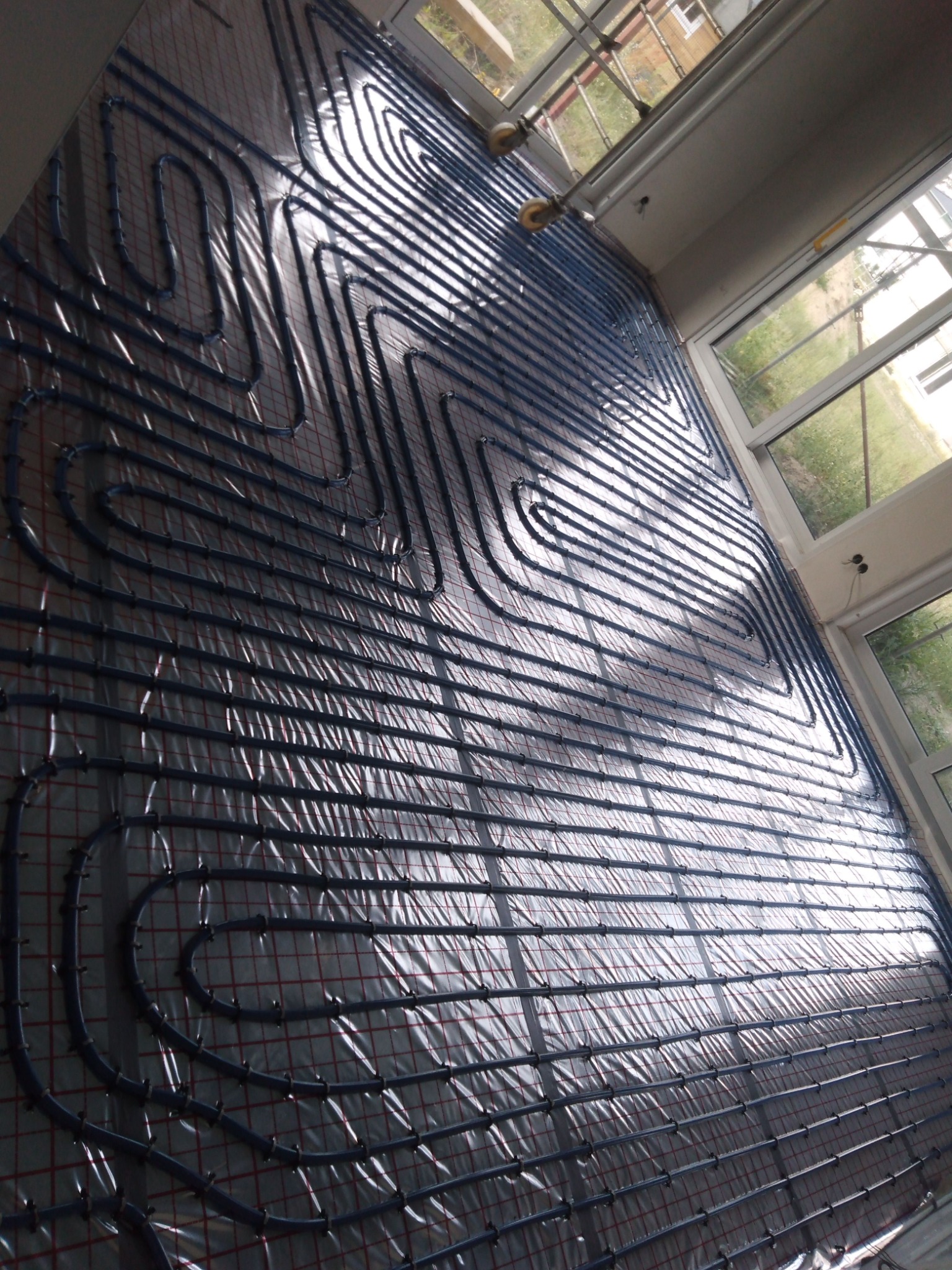 Niebieskie rury ogrzewania podłogowego ułożone na folii z czerwoną siatką, widok z góry w pomieszczeniu z oknami.