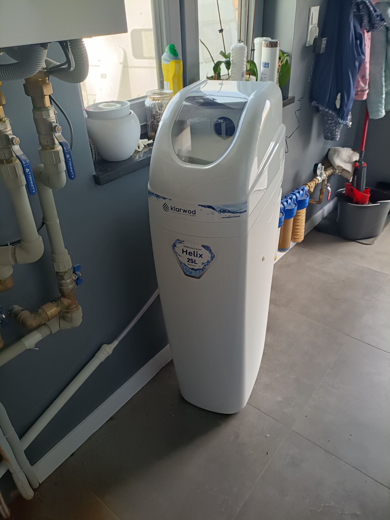 Biały zmiękczacz wody Klarwod Helix 25L, zainstalowany w pomieszczeniu z szarymi ścianami i płytkami podłogowymi, widoczne przyłącza hydrauliczne i filtry wody.