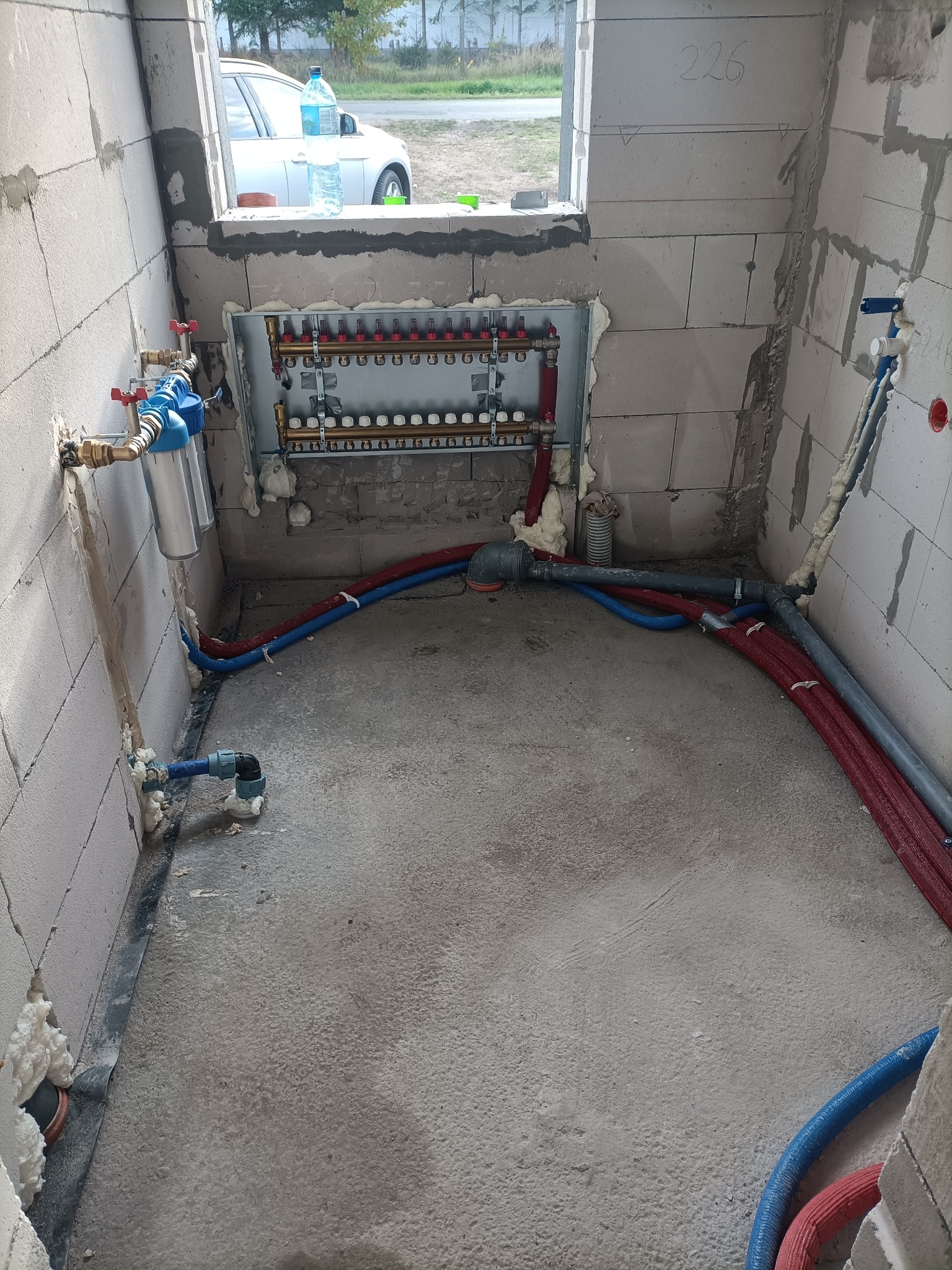 Rozdzielacz instalacji centralnego ogrzewania i wodnej, z rurami w izolacji czerwonej i niebieskiej, zamontowany w surowym pomieszczeniu z betonu komórkowego, widoczny filtr wody z niebieską obudową.