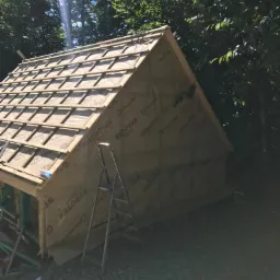 Konstrukcja dachu w trakcie budowy, widoczna membrana paroizolacyjna z nadrukiem, drewniane łaty, drabina oparta o ścianę, otoczenie leśne, słoneczny dzień.