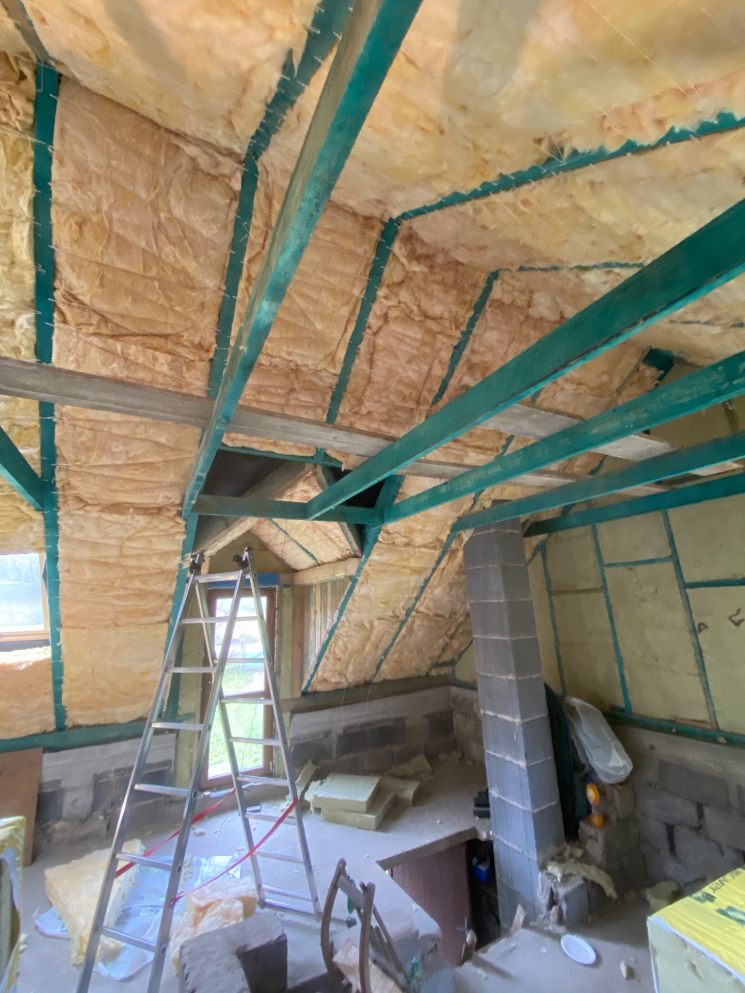 Poddasze ocieplone wełną mineralną, widoczne zielone belki konstrukcyjne, drabina aluminiowa i fragment komina z pustaków szarych.