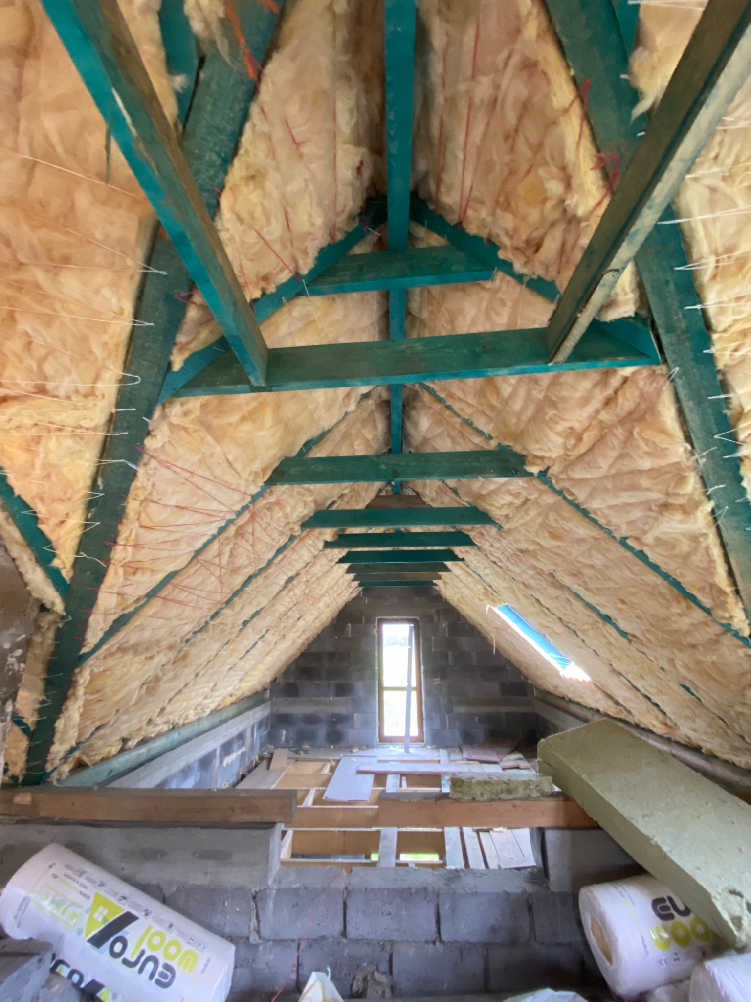 Poddasze w trakcie izolacji wełną mineralną, widoczne drewniane krokwie pomalowane na zielono, sznurki mocujące izolację oraz okno dachowe w oddali.