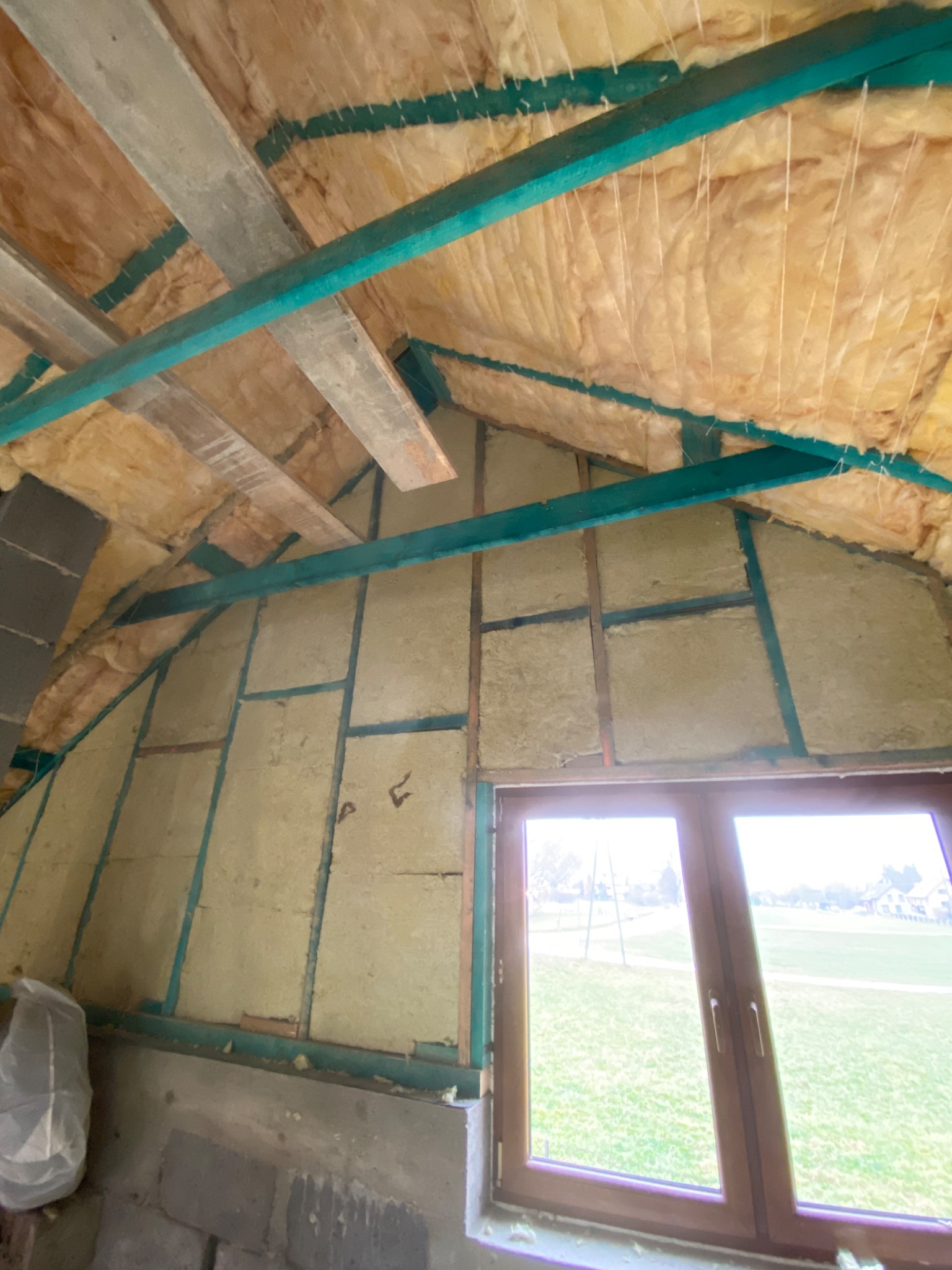 Skośne poddasze z ociepleniem z wełny mineralnej, widoczne drewniane elementy konstrukcyjne pomalowane na turkusowo i okno z widokiem na zielony trawnik.