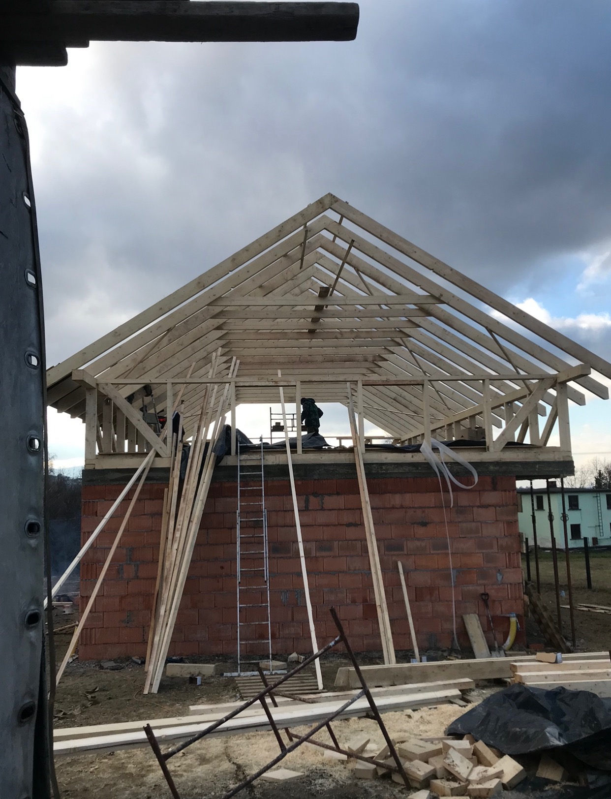Wznoszenie drewnianej konstrukcji dachu na murowanym budynku w stanie surowym otwartym, widoczna drabina i porozrzucane deski na placu budowy, pochmurne niebo w tle.