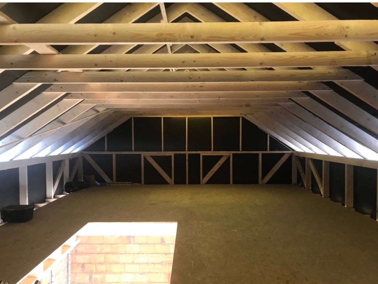 Poddasze w trakcie adaptacji: widoczne drewniane belki stropowe, czarna izolacja na ścianach szczytowych, oraz przygotowana podłoga. W tle narzędzia i wiadro.