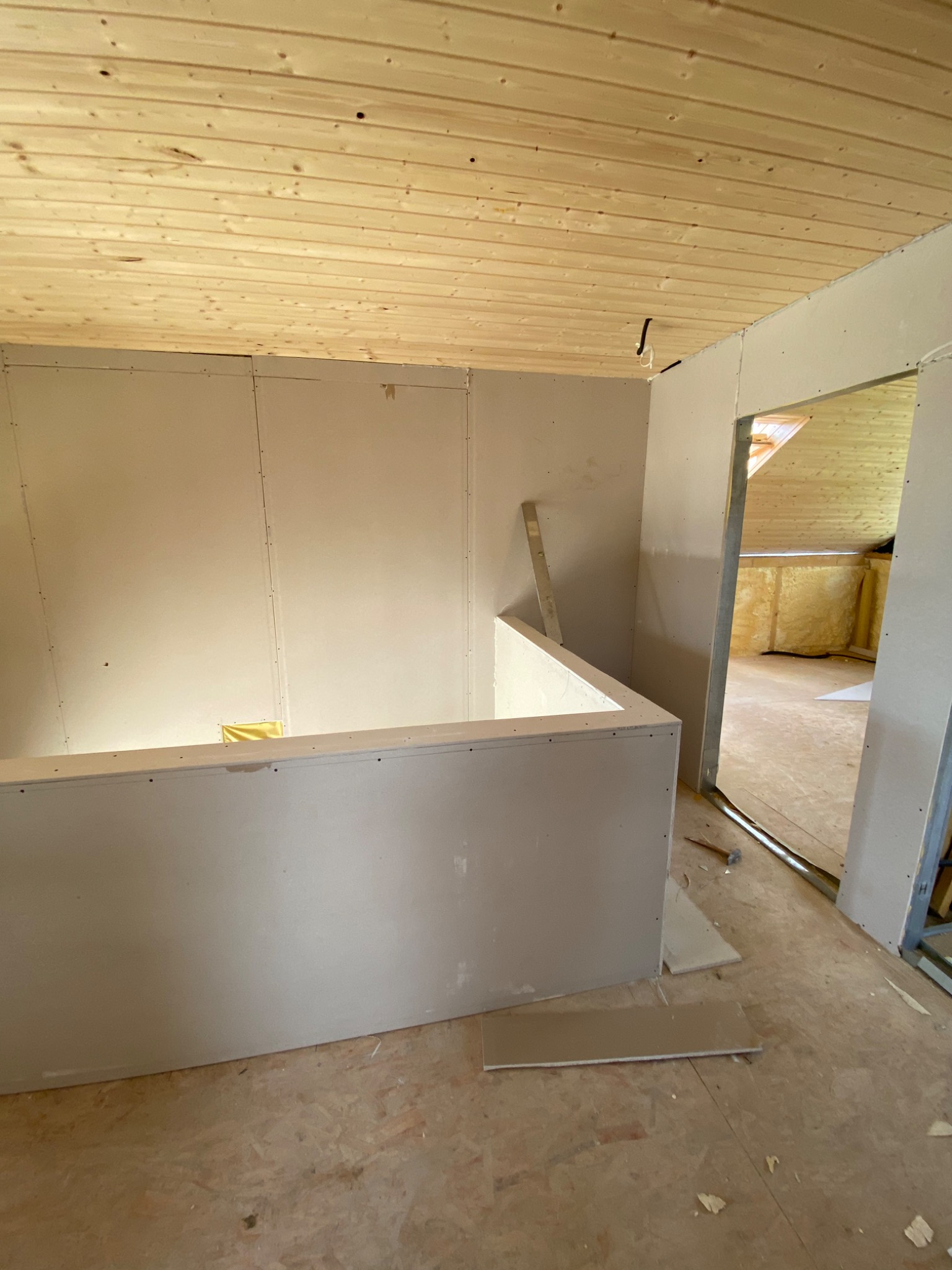 Wnętrze poddasza w trakcie wykańczania płytami kartonowo-gipsowymi, widoczna drewniana konstrukcja sufitu i ocieplenie w sąsiednim pomieszczeniu. Częściowo zamontowana ścianka działowa z płyt G-K.