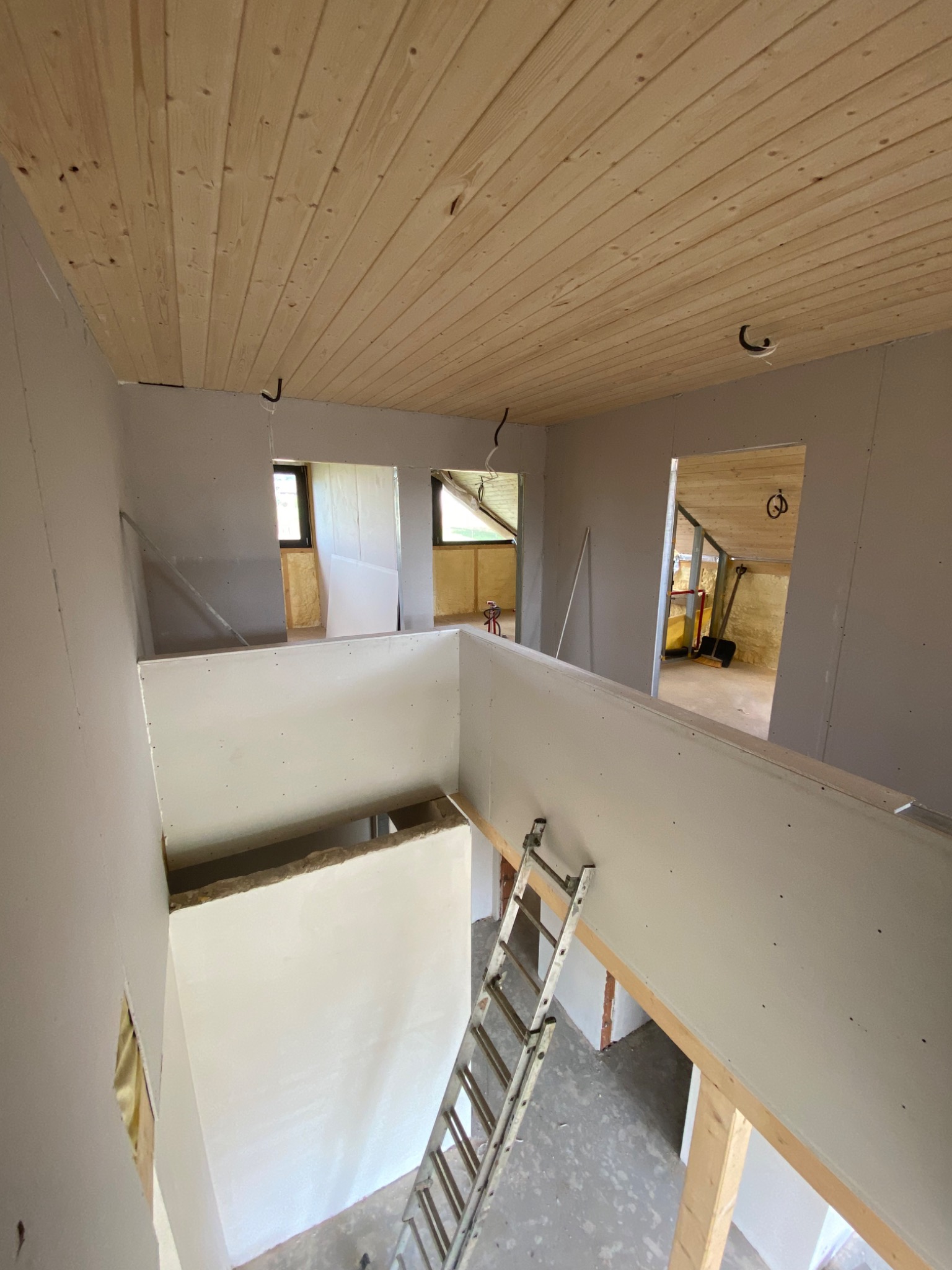 Poddasze w trakcie wykańczania: drewniana boazeria na suficie, ściany z płyt kartonowo-gipsowych, widoczne otwory okienne i drabina oparta o ścianę.