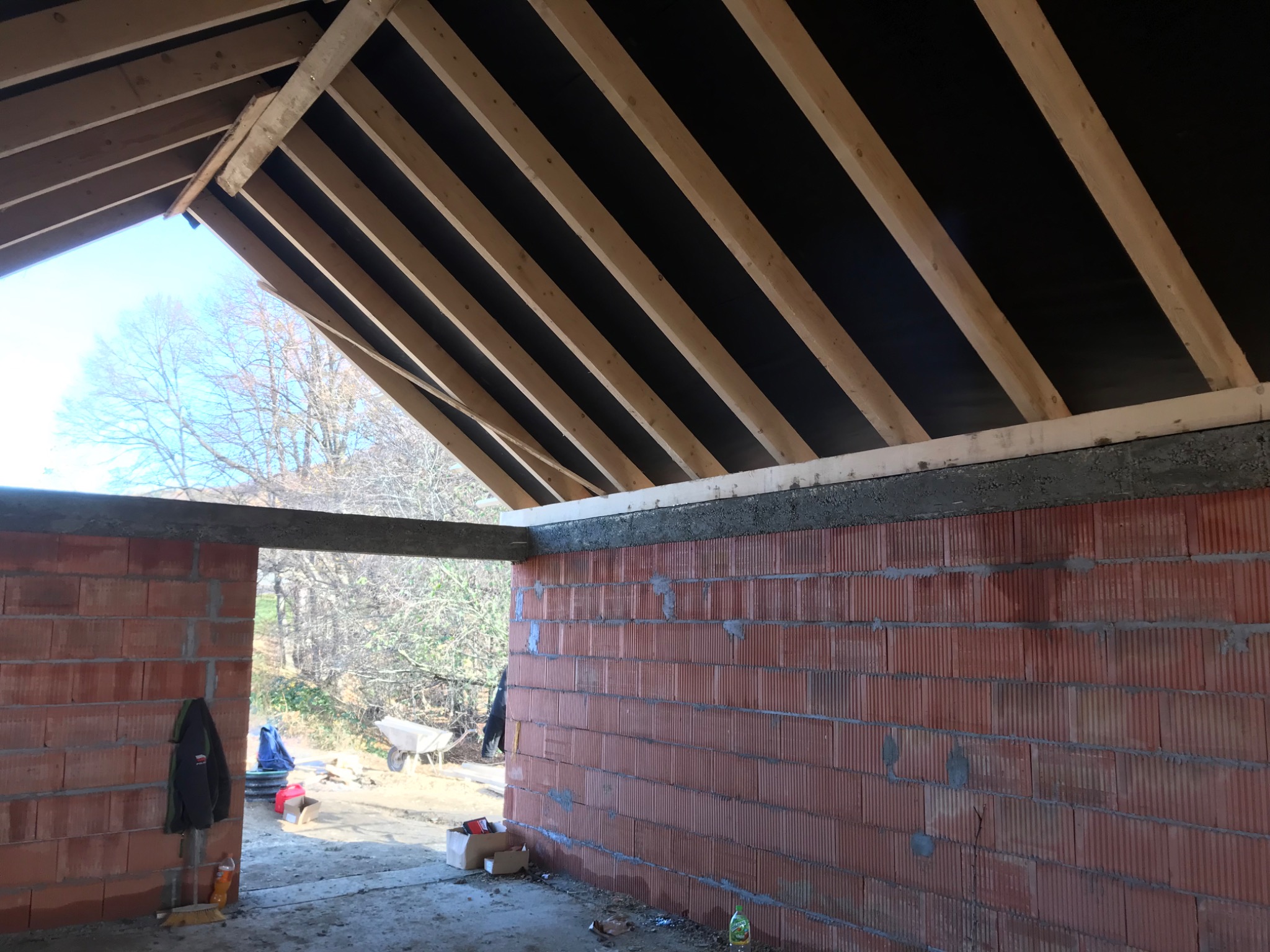 Surowy stan poddasza z widoczną konstrukcją dachu, ociepleniem wstępnym i murowanymi ścianami z pustaków ceramicznych, widok przez otwór drzwiowy na zewnątrz budynku.