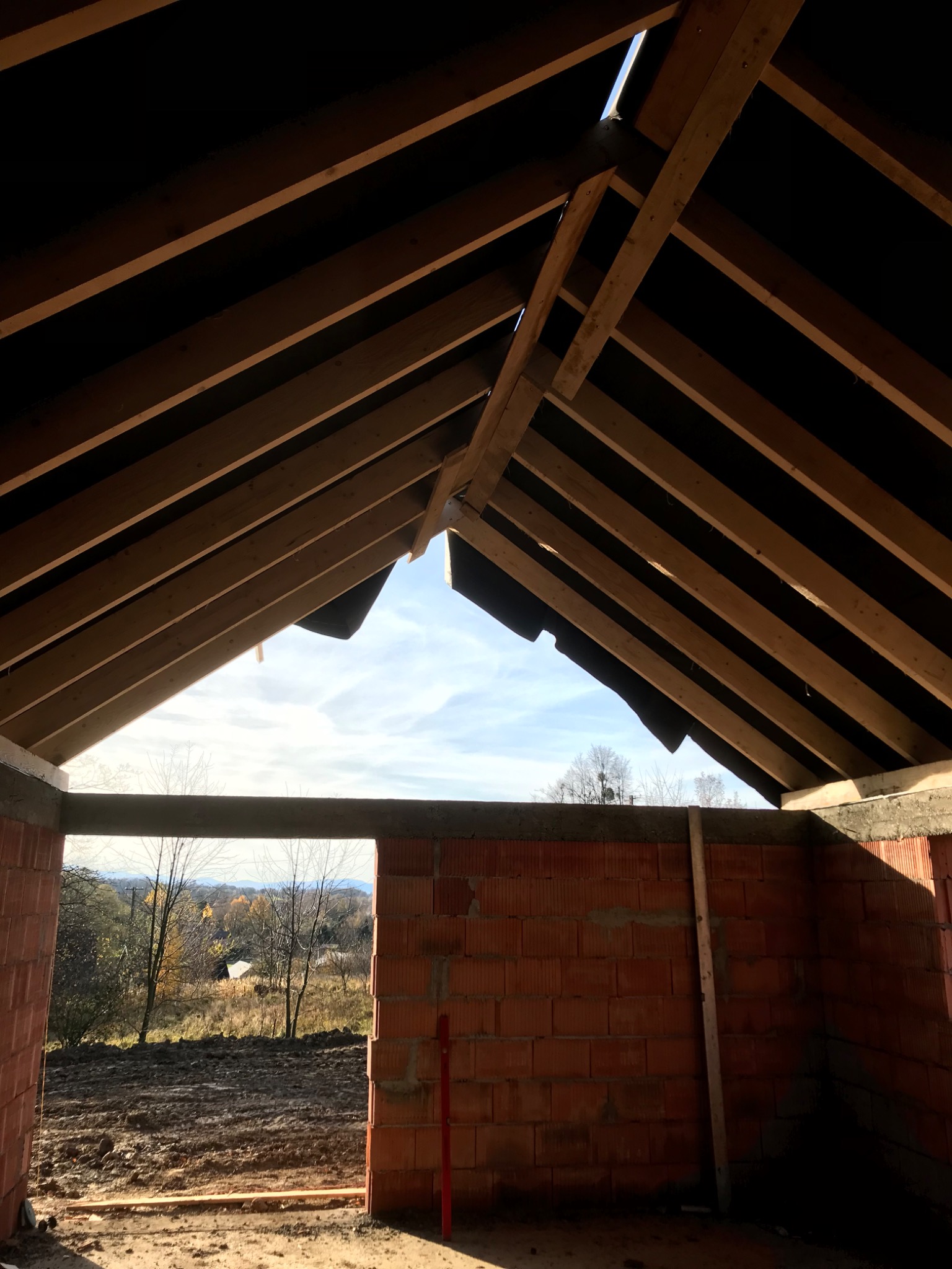 Wnętrze surowego domu w trakcie budowy, widoczne drewniane krokwie dachu i ściany z czerwonej cegły, w tle jesienny krajobraz za oknem.