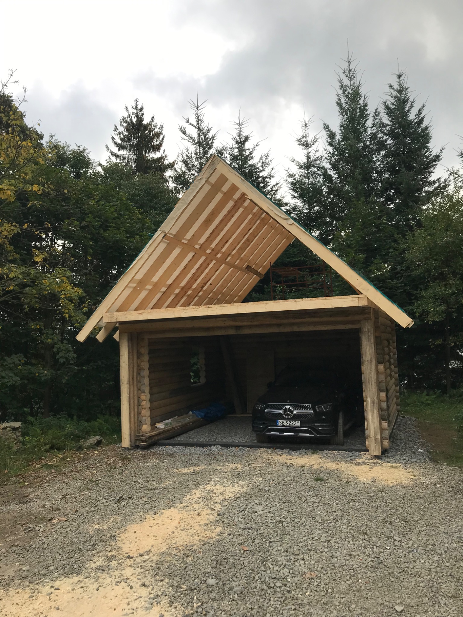 Surowy, drewniany garaż z bali w trakcie budowy, z częściowo wykończonym dachem i zaparkowanym czarnym samochodem w środku, otoczony drzewami.