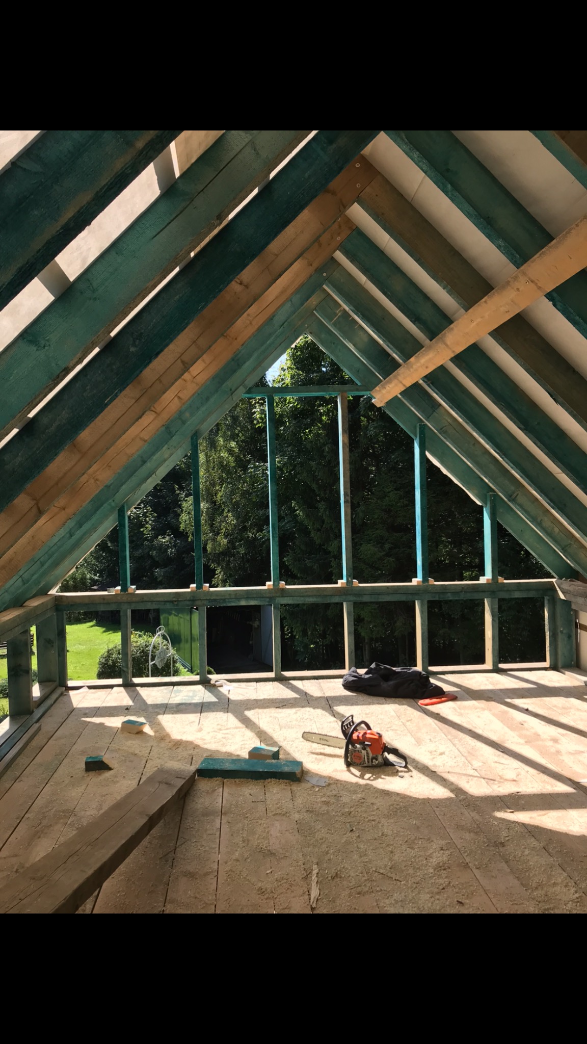 Konstrukcja drewniana poddasza w trakcie budowy, widoczne krokwie pomalowane na zielono, deski podłogowe pokryte pyłem po cięciu, piła spalinowa Stihl leżąca na podłodze, widok na zieleń za oknem.