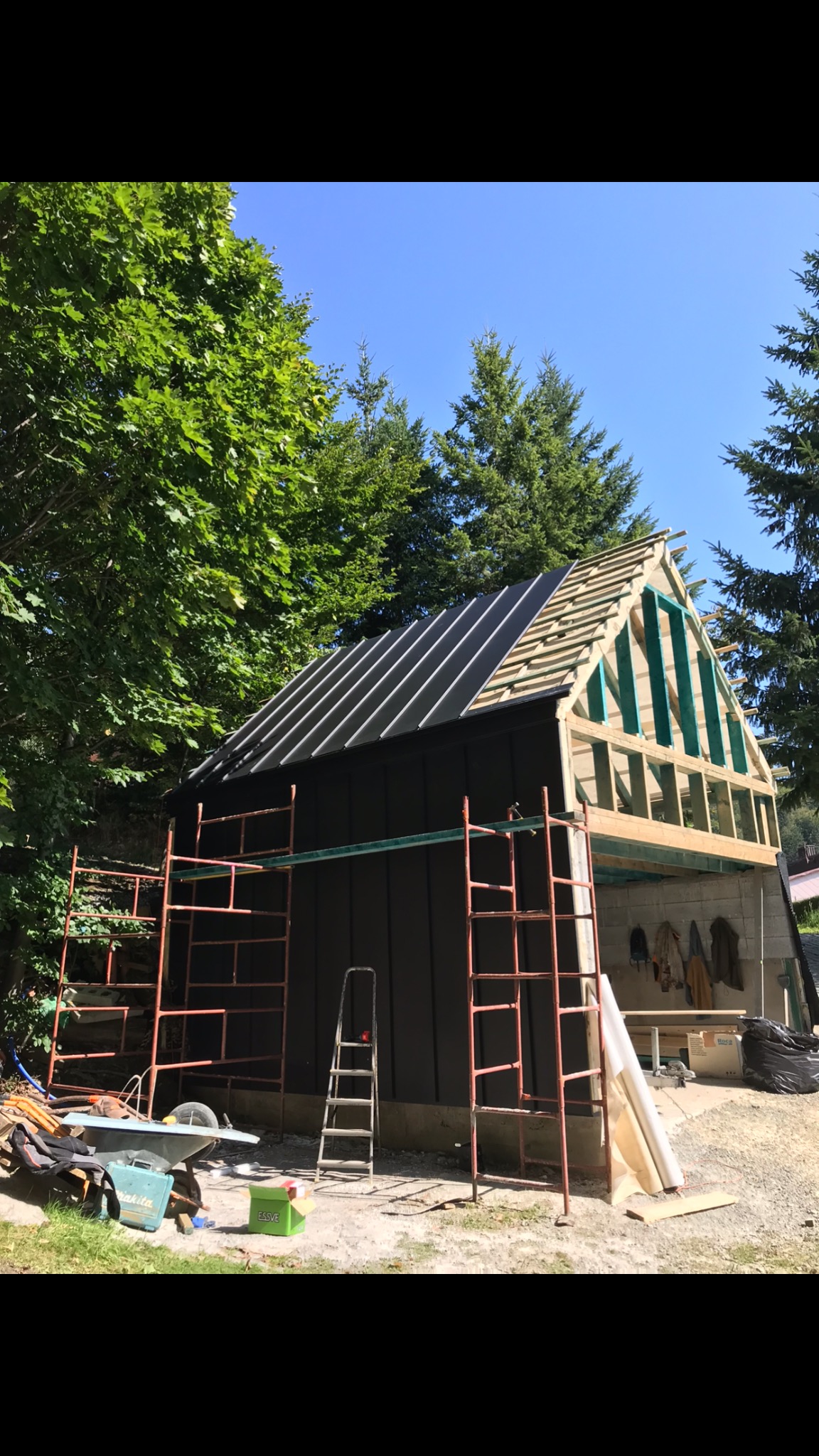 Budowa szkieletu budynku z widocznym ociepleniem dachu, rusztowaniem i narzędziami na zewnątrz, w tle drzewa i błękitne niebo.