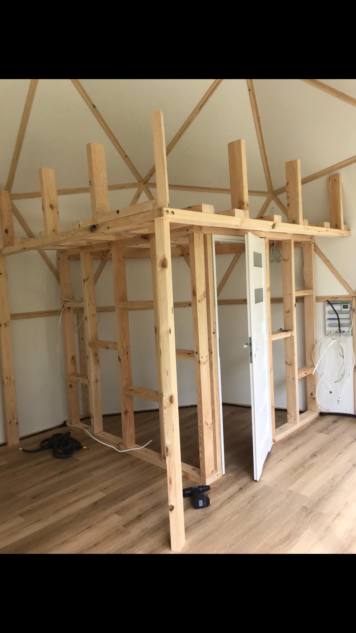 Wnętrze domu w trakcie budowy z drewnianą konstrukcją antresoli, widoczne instalacje elektryczne, podłoga z paneli oraz białe drzwi.