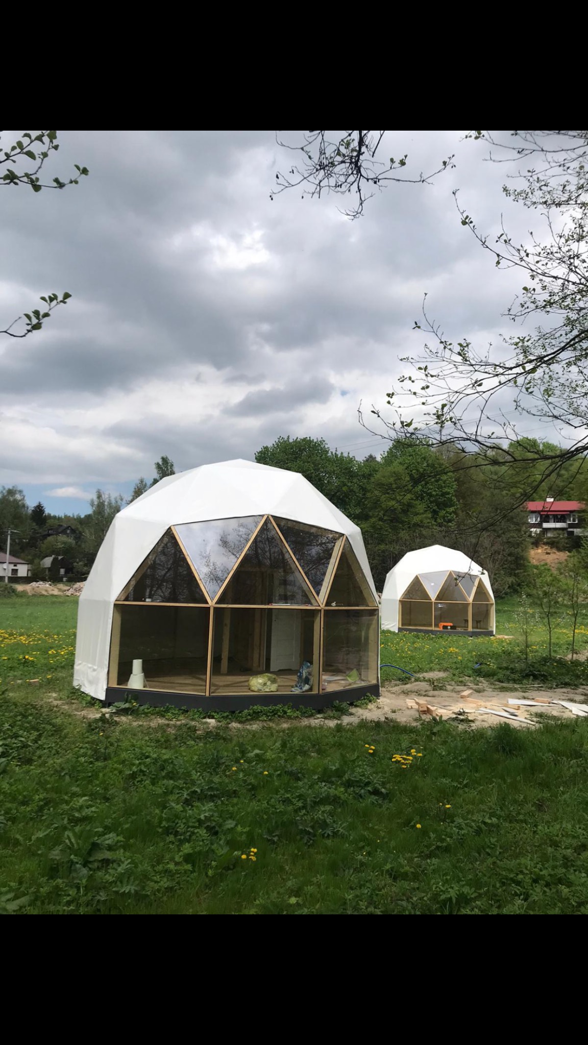 Dwa białe geodezyjne domy kopułowe z drewnianą ramą i szklanymi panelami na zielonej łące z drzewami w tle, widoczne ślady budowy.