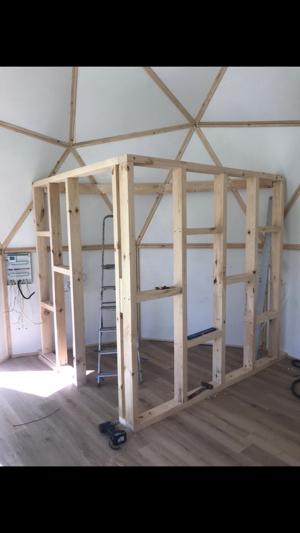 Wnętrze kopuły geodezyjnej w trakcie budowy ścian działowych z drewnianej ramy, widoczna drabina, wiertarko-wkrętarka akumulatorowa i panel elektryczny na ścianie.