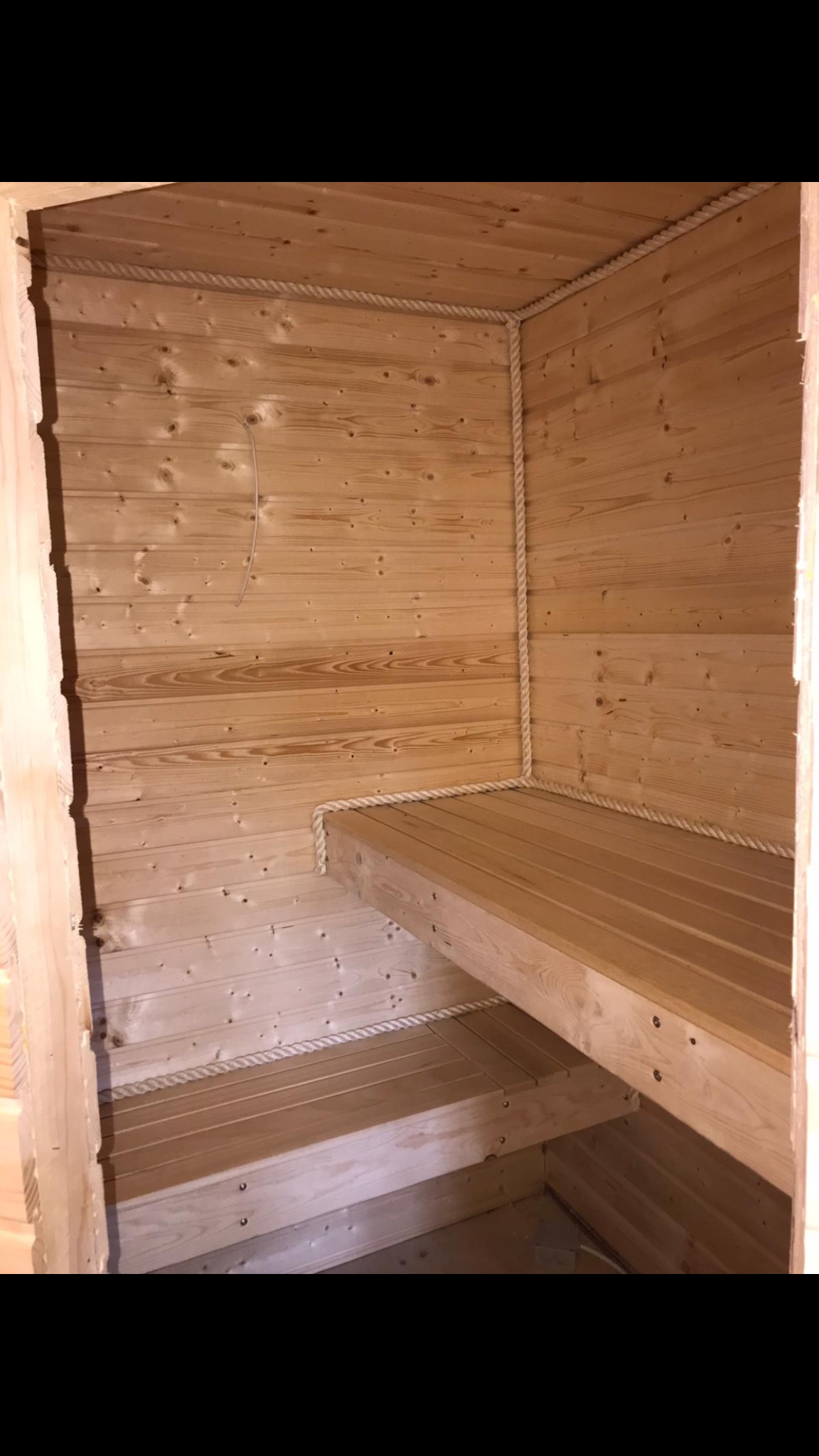 Wnętrze sauny z jasnego drewna wykończone grubym, białym sznurem, widoczne ławki i surowe ściany z desek.