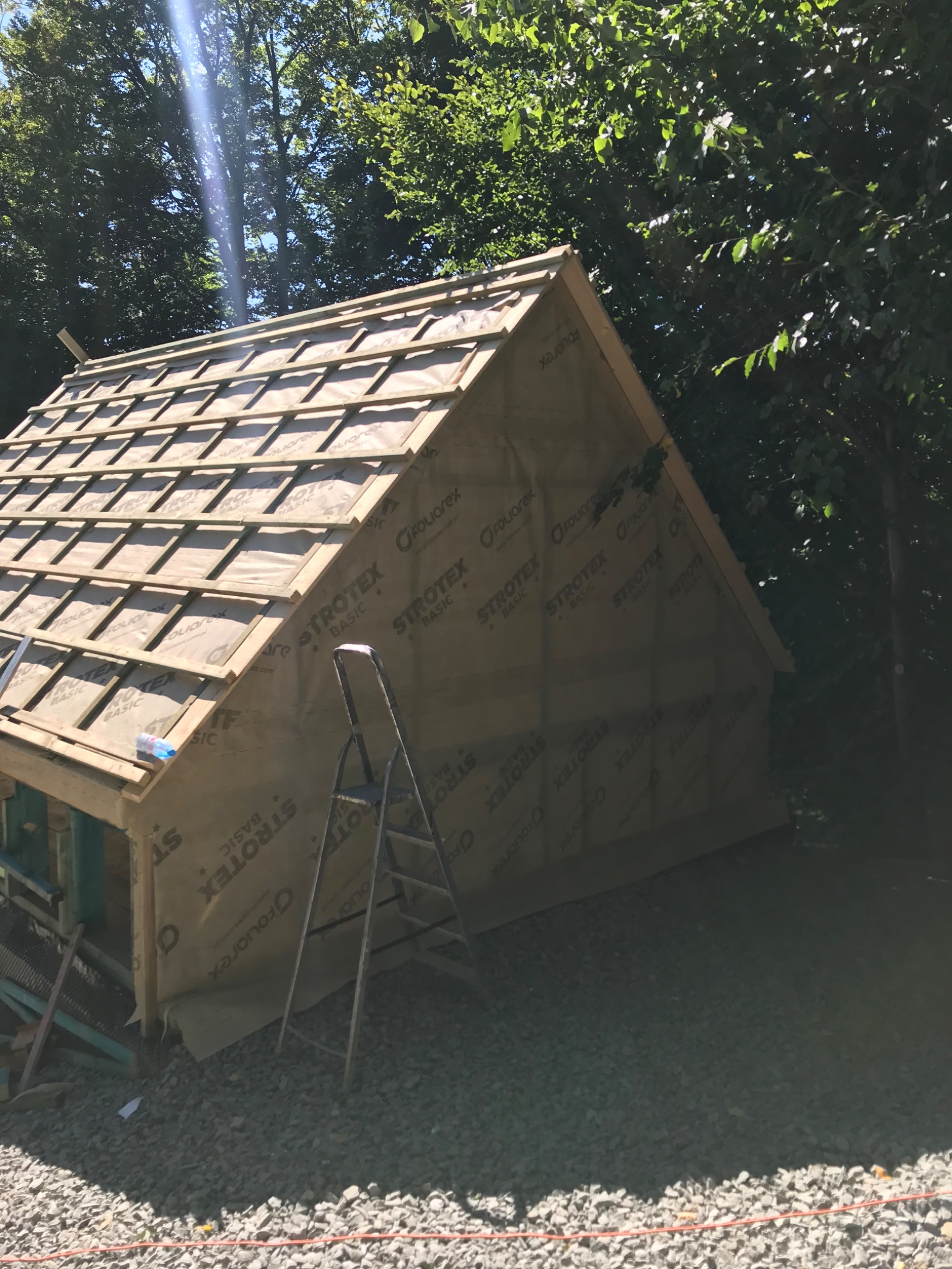 Konstrukcja dachu w trakcie budowy, widoczna membrana paroizolacyjna z nadrukiem, drewniane łaty, drabina oparta o ścianę, otoczenie leśne, słoneczny dzień.