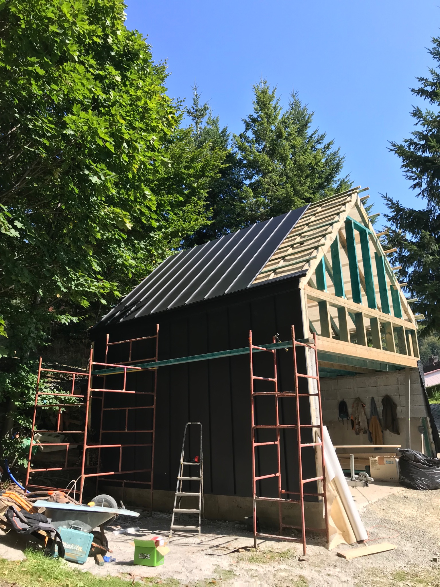 Budynek w trakcie budowy z widoczną konstrukcją dachu i częściowo położonym czarnym pokryciem, rusztowanie i drabina oparta o ścianę, otoczenie z zielenią drzew.