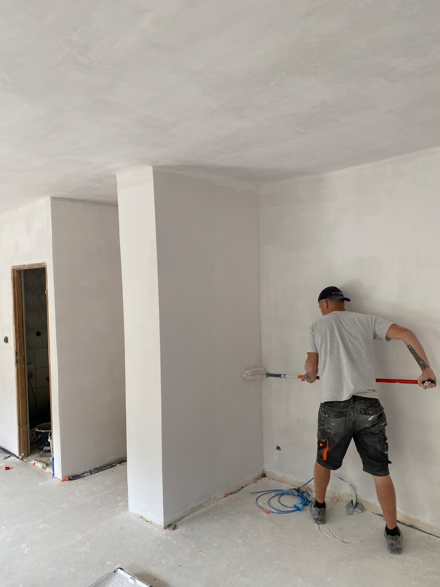 Malarz w szortach i czapce maluje ścianę wałkiem w jasnym, surowym wnętrzu w trakcie remontu; widoczne kable i narzędzia malarskie.