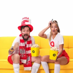 Para kibiców w barwach Polski, siedząca na żółtej kanapie z popcornem, z zaskoczonymi minami ogląda mecz, kobieta trzyma pilot w ręce.