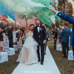 www.fotokutkowska.pl - Para młoda w trakcie ceremonii ślubnej, otoczona gośćmi z kolorowymi racami dymnymi na tle zieleni parku.