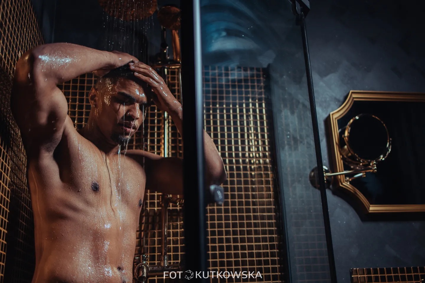 Mężczyzna pod prysznicem z złotą mozaiką w tle, woda spływa po jego ciele, w tle lustro w złotej ramie.