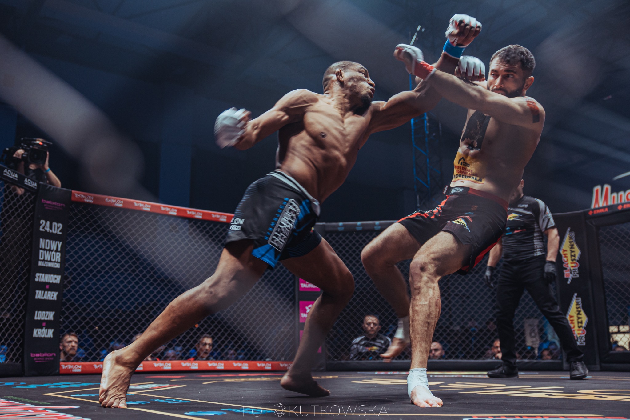 Dwóch zawodników MMA w dynamicznej wymianie ciosów na ringu, jeden z nich w czarnych spodenkach z niebieskimi akcentami, drugi w czarno-czerwonych, w tle siatka ringu, publiczność i oświetlenie hali.