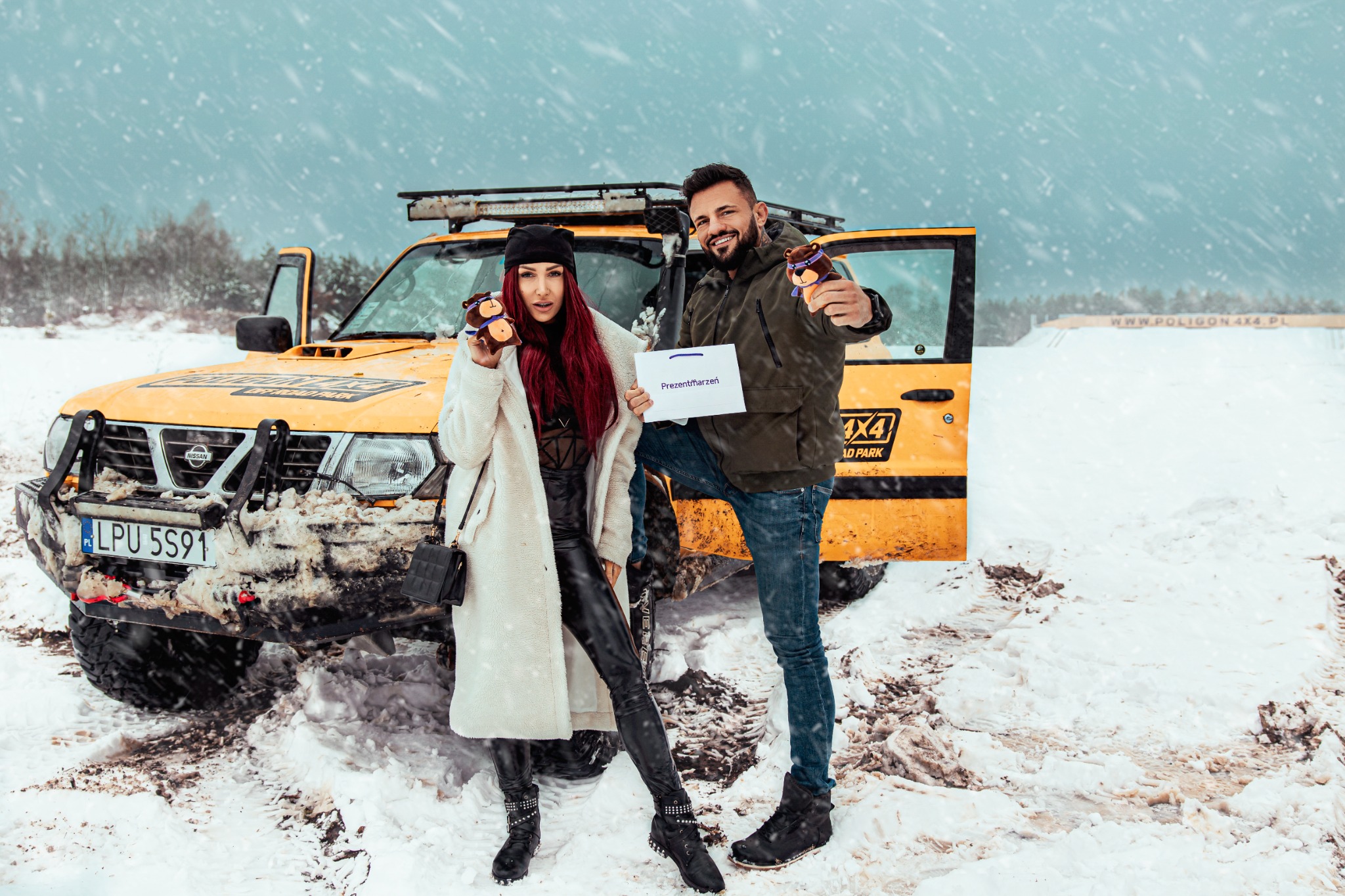 Para trzymająca pluszowe misie i kartkę z napisem 'PrezentMarzeń' na tle żółtego samochodu terenowego 4x4 w zimowej scenerii podczas opadów śniegu.