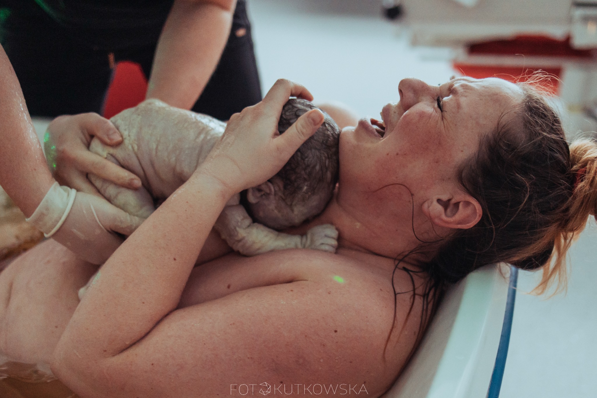 Emocjonalne ujęcie z porodu w wodzie: kobieta z zamkniętymi oczami i otwartymi ustami trzyma noworodka, wspierana przez osobę w rękawiczkach; w tle widoczny fragment wanny.