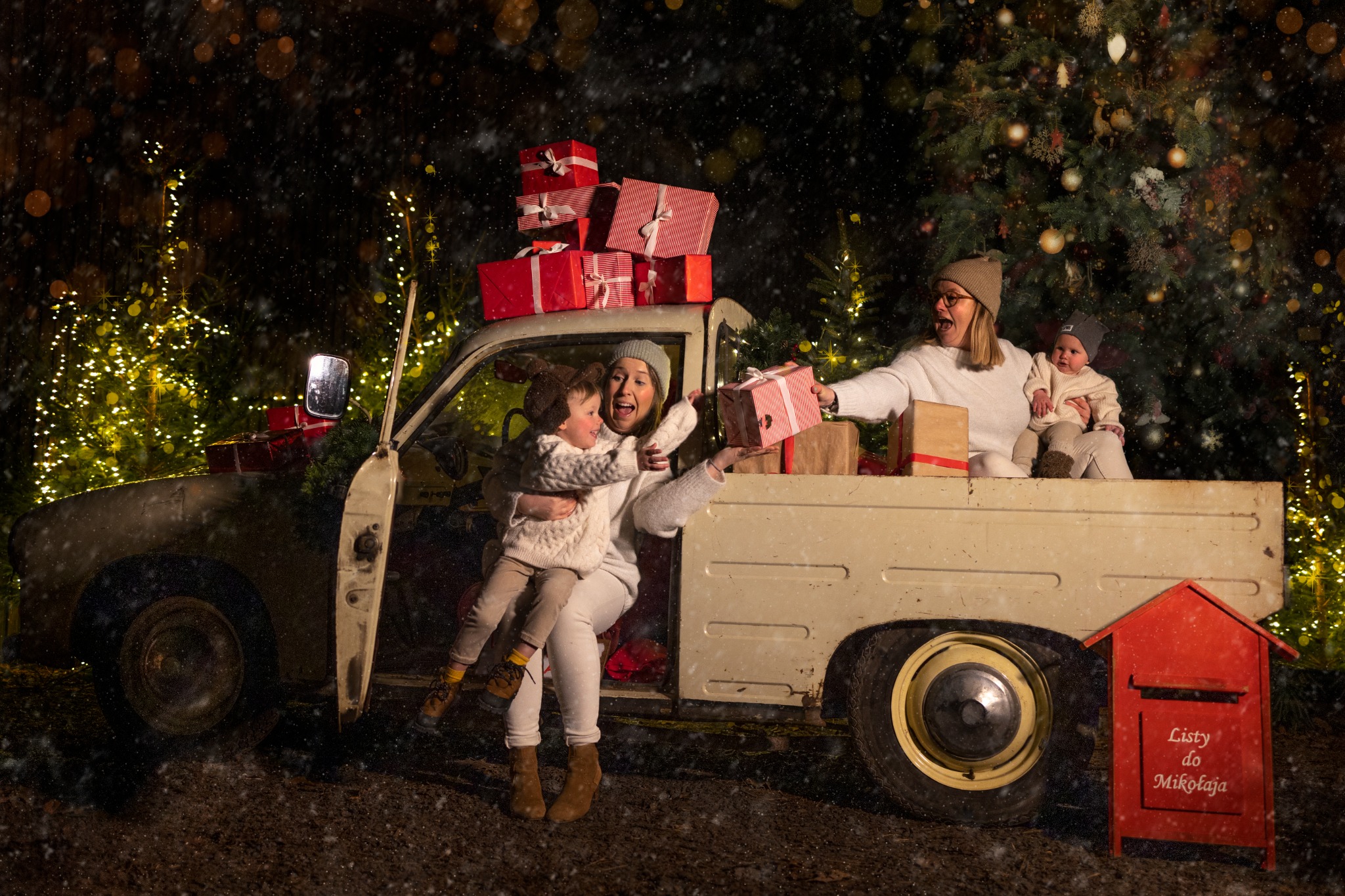 Świąteczna sesja rodzinna przy starym samochodzie z prezentami i skrzynką na listy do Mikołaja, w tle oświetlone choinki i padający śnieg.