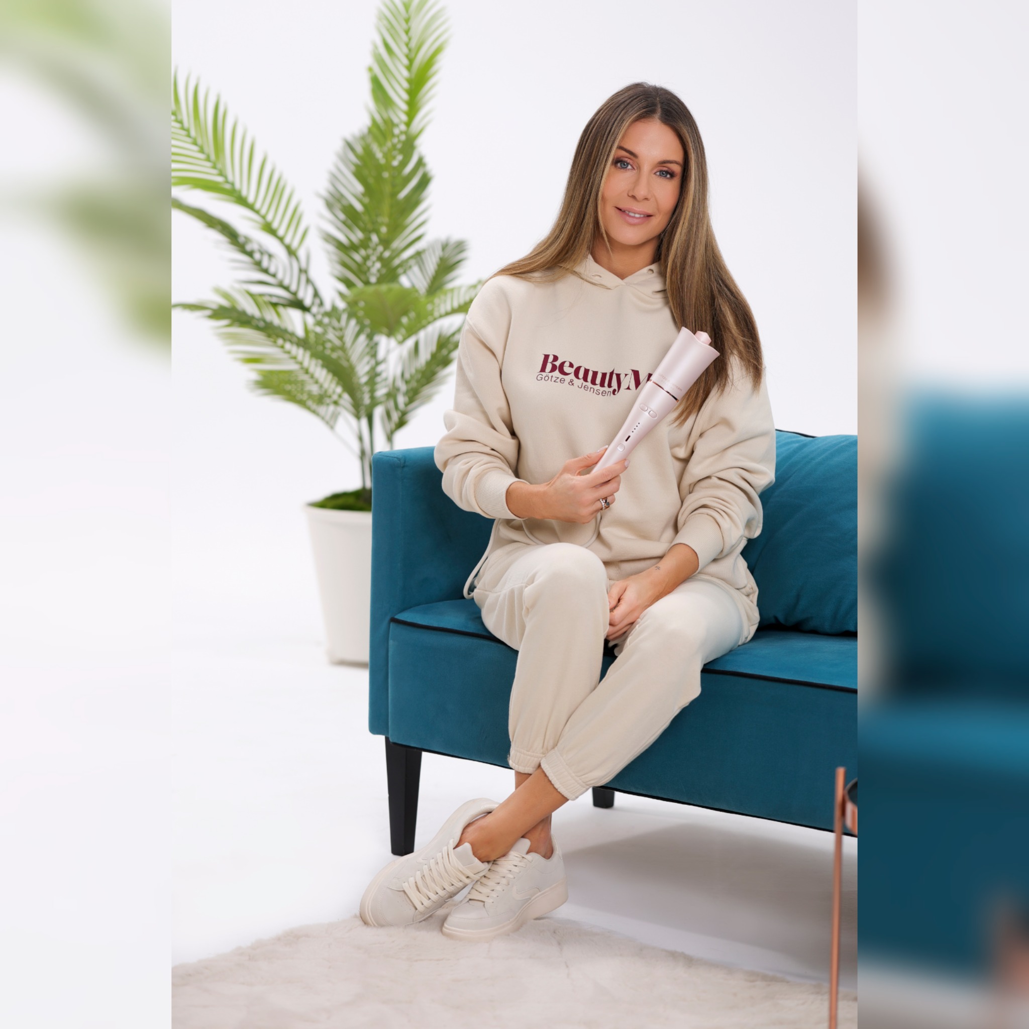 Kobieta siedzi na turkusowej sofie, trzymając w dłoniach różową lokówkę, ubrana w beżowy dres z logo, obok doniczkowa palma.