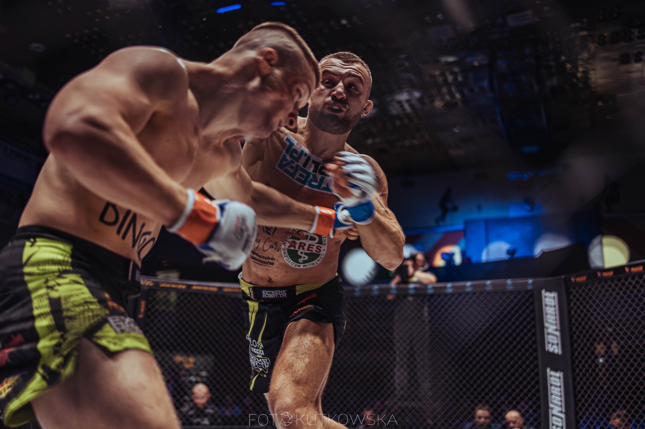 Dwóch zawodników MMA w trakcie walki w klatce, jeden z nich z tatuażami i grymasem na twarzy, w tle widownia.