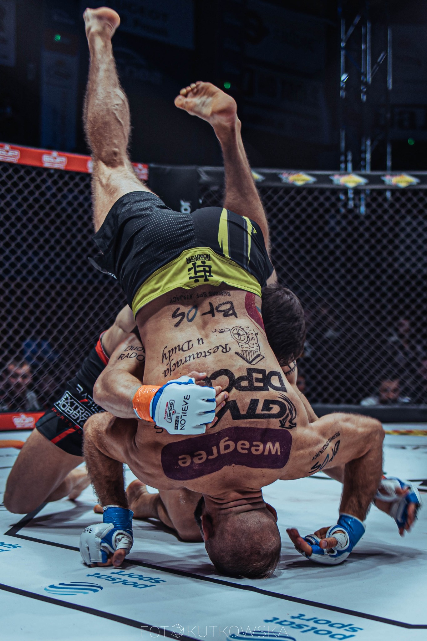 Dwóch zawodników MMA w trakcie walki: jeden trzyma drugiego do góry nogami w klatce, widoczne tatuaże na plecach i ramionach zawodników.