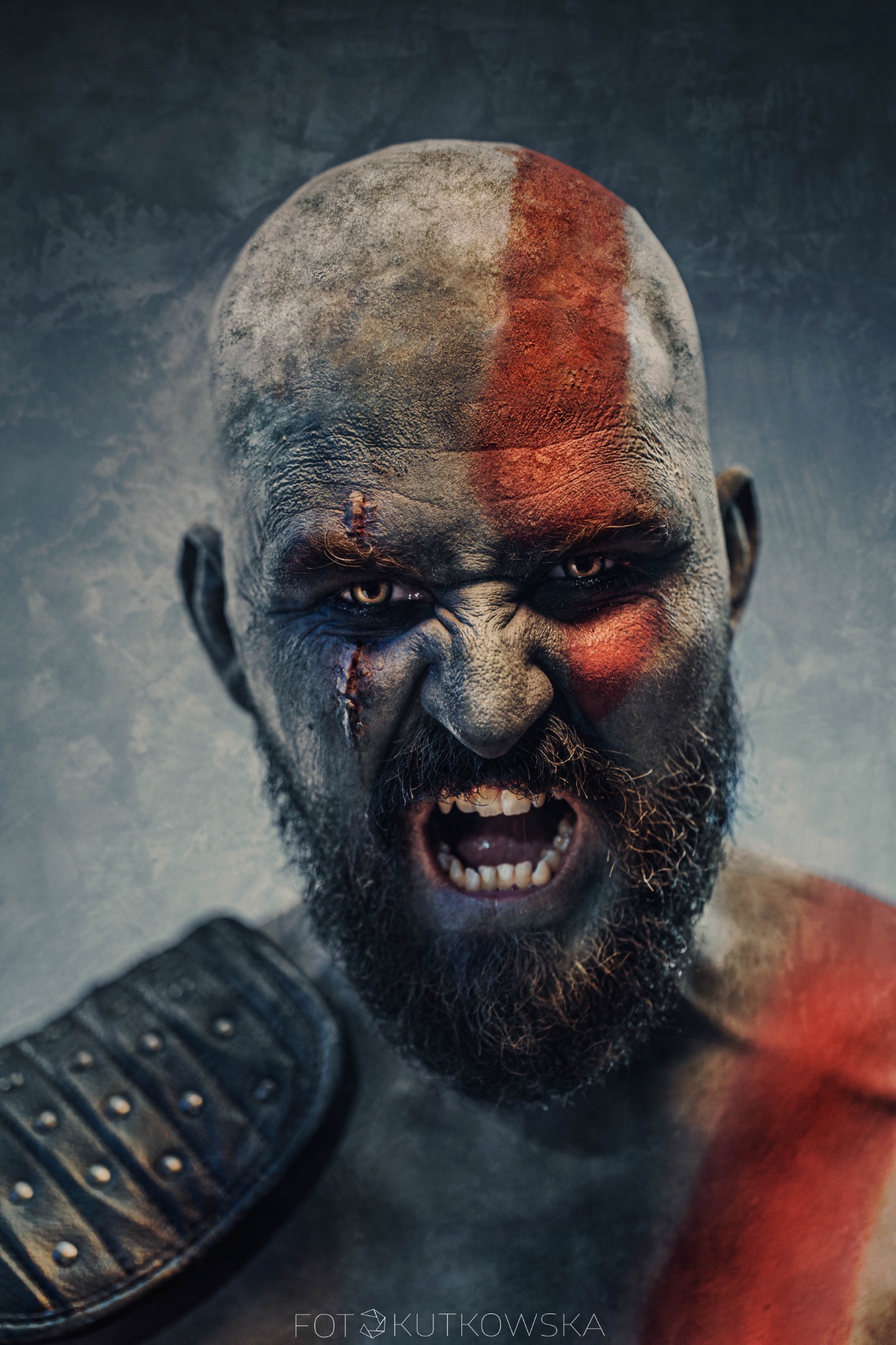 Mężczyzna z pomalowaną twarzą i brodą, stylizowany na postać z gry, z widocznymi bliznami i grymasem na twarzy, ubrany w elementy zbroi na ramieniu.
