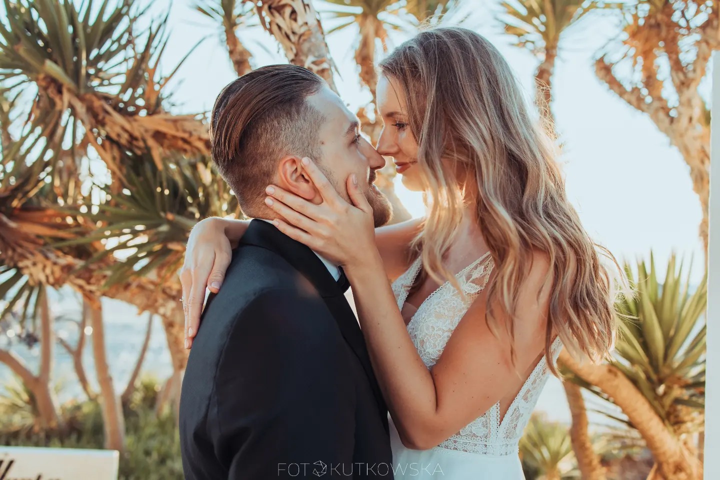 Uśmiechnięta para, panna młoda w koronkowej sukni, obejmuje pana młodego na tle egzotycznych drzew w słoneczny dzień.