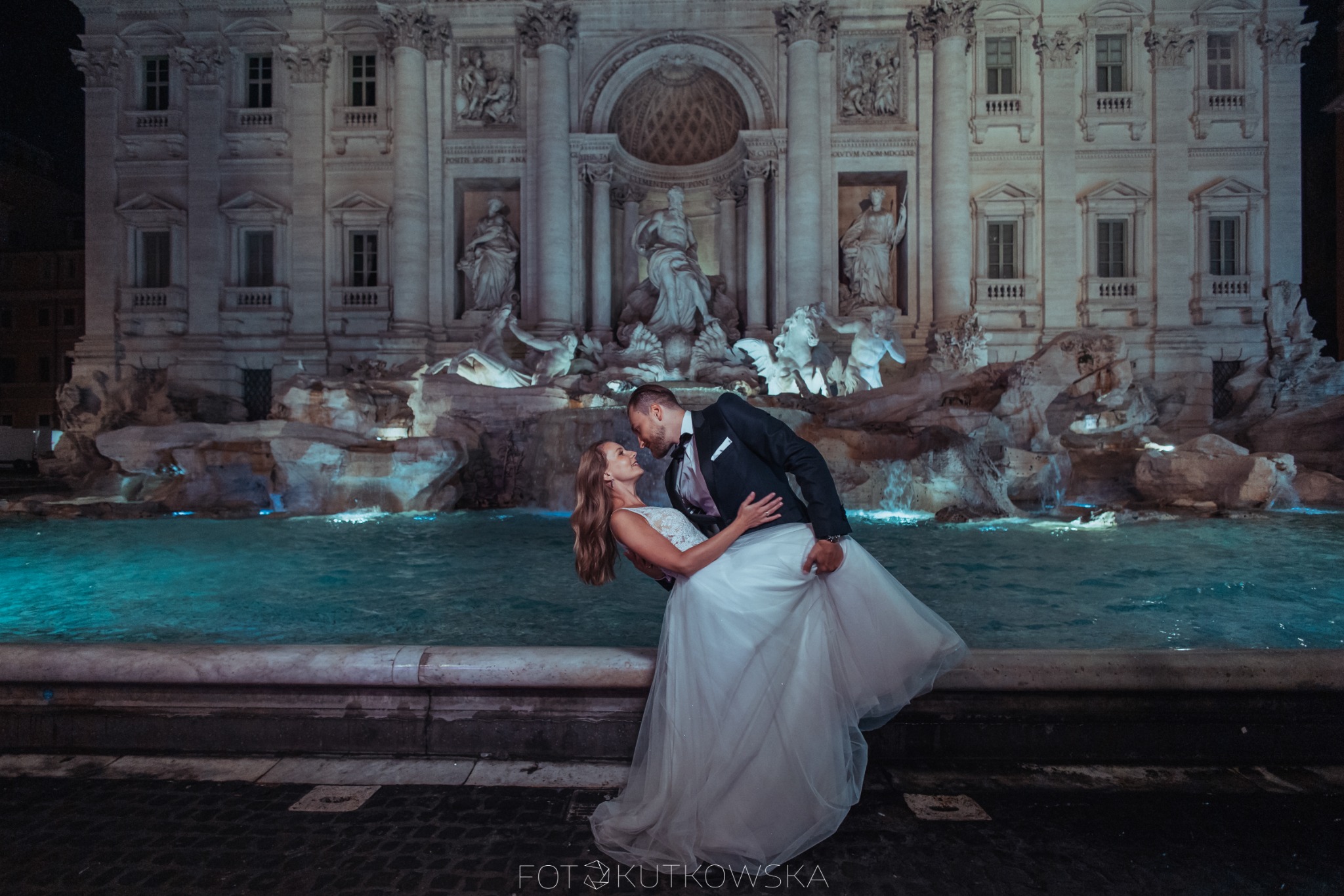 Para młoda w tańcu przed oświetloną Fontanną di Trevi w Rzymie, panna młoda w białej sukni z koronkową górą, pan młody w czarnym smokingu z muszką.