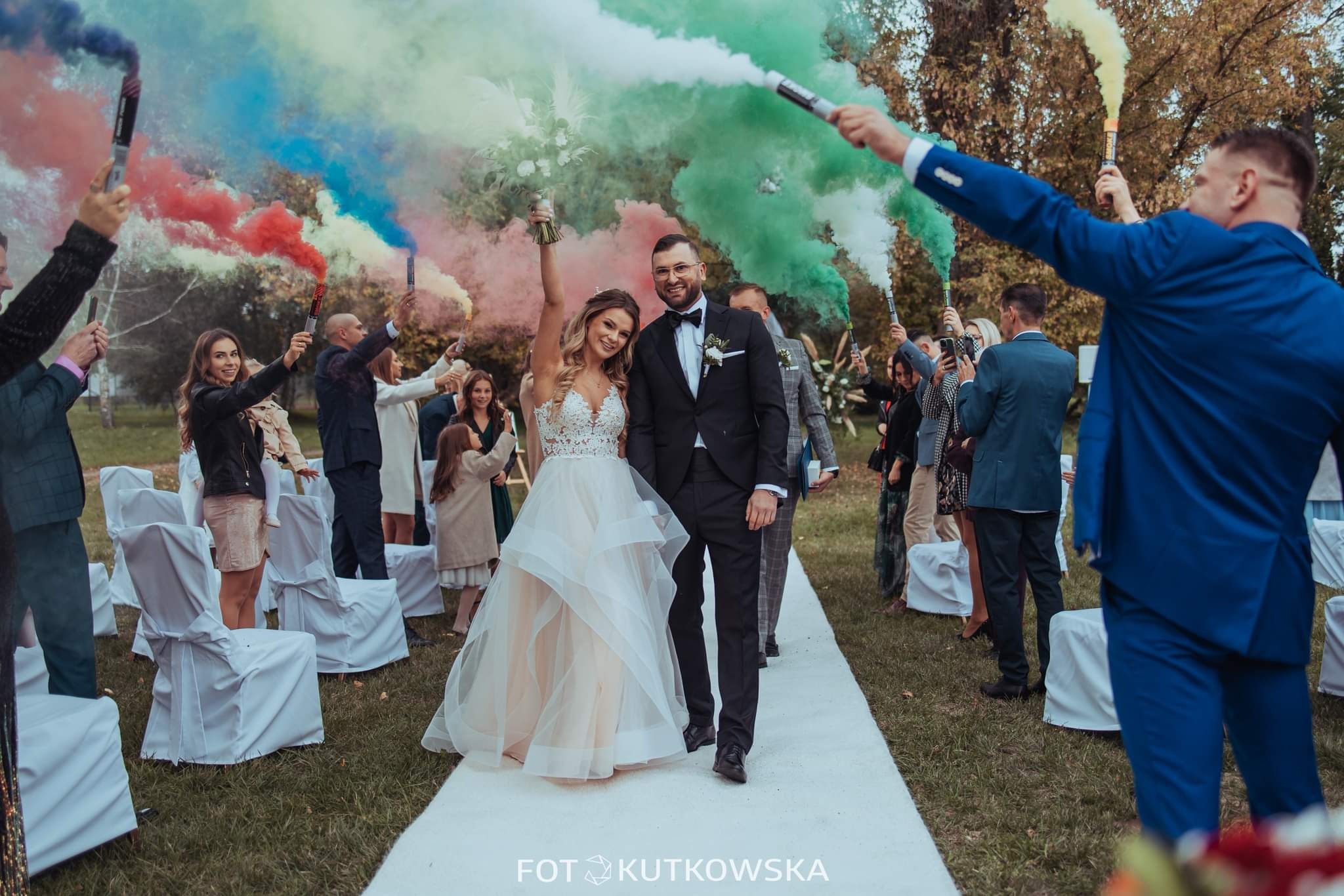 Para młoda w trakcie ceremonii ślubnej, otoczona gośćmi z kolorowymi racami dymnymi na tle zieleni parku.