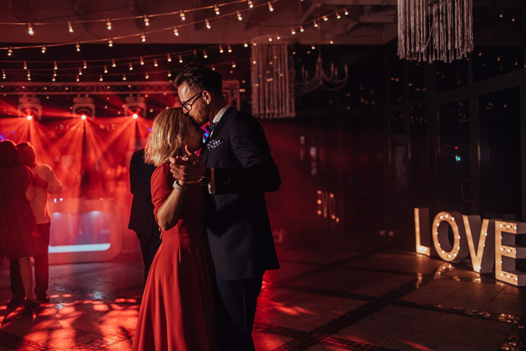 Para w tańcu, mężczyzna w granatowym garniturze całuje kobietę w czerwonej sukience w czoło, dekoracje świetlne w tle, napis LOVE z podświetlanych liter.