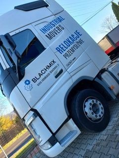 Ciężarówka z napisem 'Industrial Movers. Relokacje Przemysłowe', zaparkowana na brukowanej powierzchni. Widoczny fragment górzystego krajobrazu w tle.