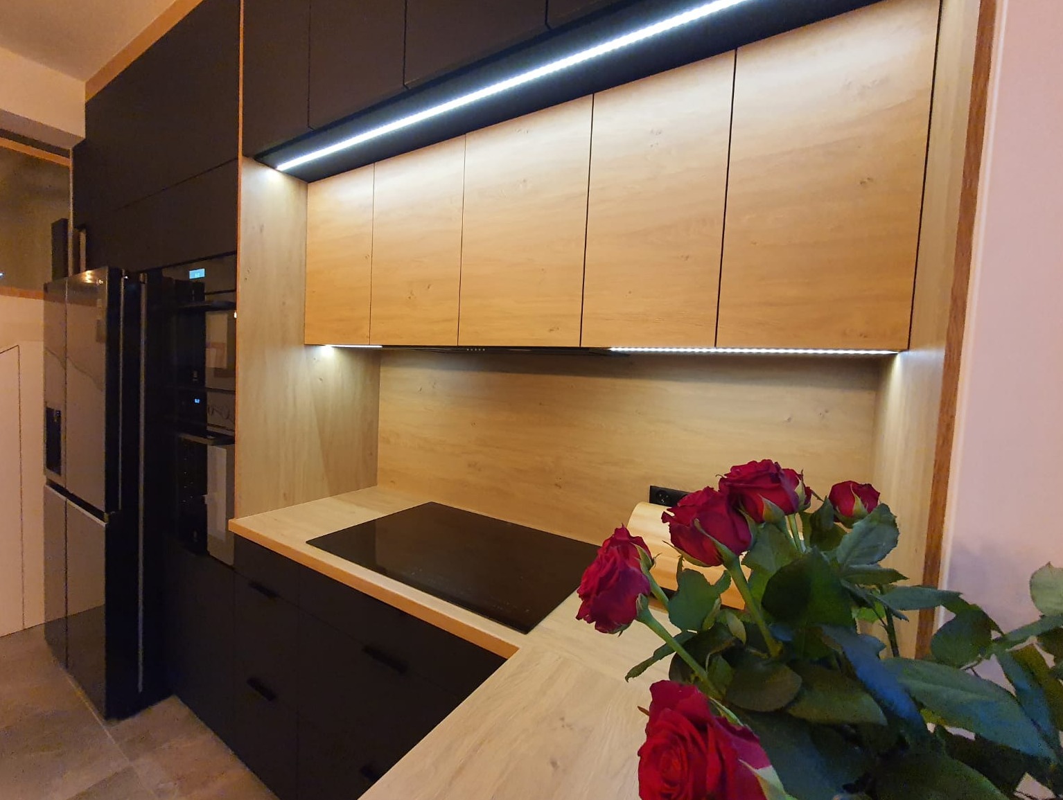 Nowoczesna kuchnia z drewnianymi szafkami górnymi podświetlonymi LED i czarnymi szafkami dolnymi, z blatem z jasnego drewna i bukietem czerwonych róż na pierwszym planie.
